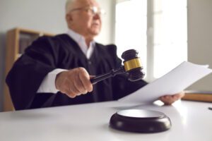 an old man with glasses wearing a judge's black robe is holding a document, possibly a court order, in one hand and swinging a gavel down with his other hand, signifying that he is entering an official court order in a legal case