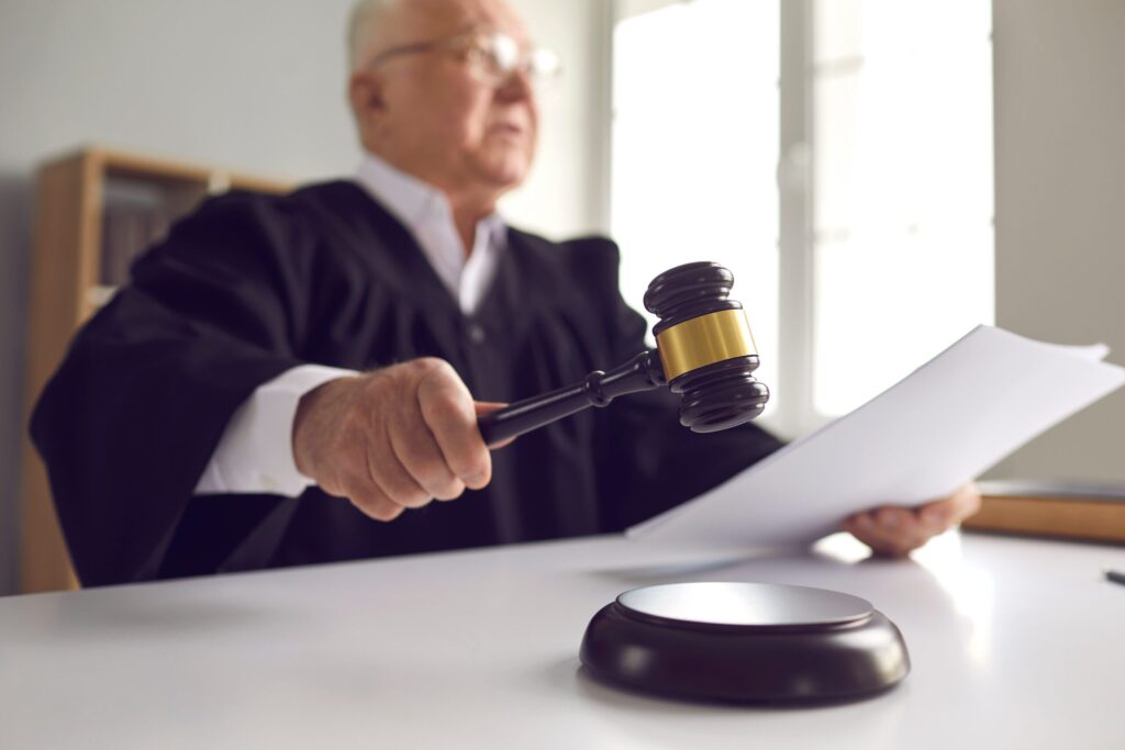 An old man with glasses wearing a judge's black robe is holding a document, possibly a court order, in one hand and swinging a gavel down with his other hand, signifying that he is entering an official court order in a legal case.