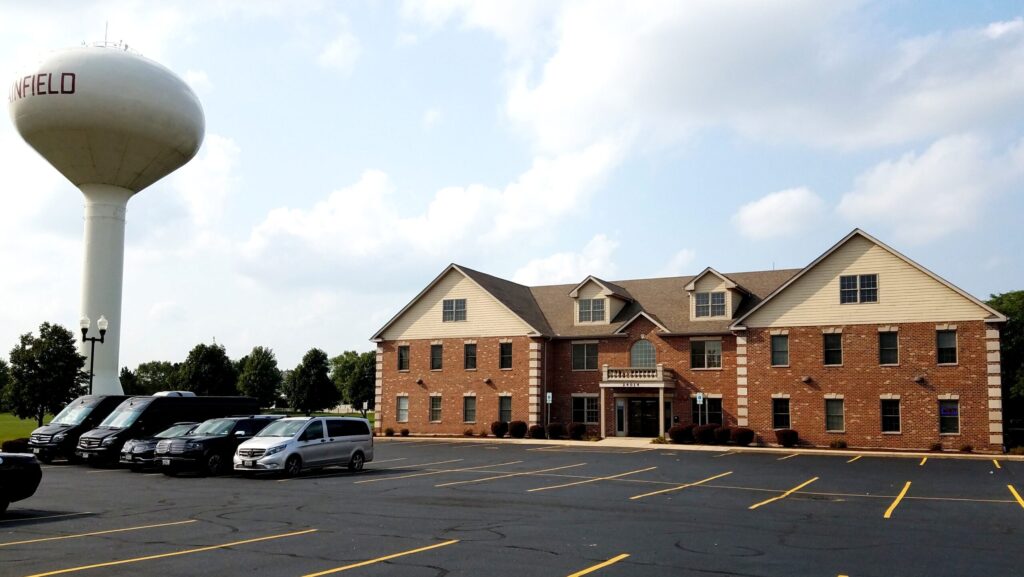 A two-story brick building with multiple windows, containing a custody attorney office building, is located in a parking lot. A large water tower labeled 'Plainfield' stands nearby under a partly cloudy sky. Several vehicles are parked in the lot.
